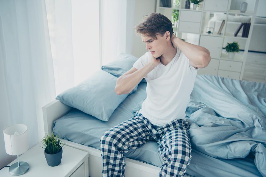 Junger Mann sitzt auf Bett mit Nackenschmerzen - Neue Matratze: Eingewöhnungszeit und körperliche Umstellung