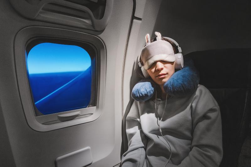 Frau schläft im Flugzeug mit Nackenhörnchen, Kopfhörern, und Schlafmaske