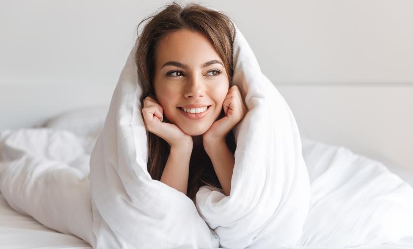 Eine junge Frau hat sich in ihre Bettdecke eingekuschelt und lächelt glücklich
