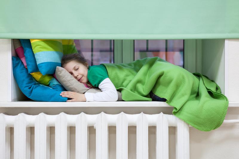 Ein kleines Mädchen hat es sich mit Bettdecke und Kissen auf dem Fensterbrett über dem Heizkörper gemütlich gemacht