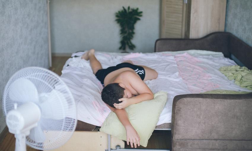 Junger Mann liegt im Bett, Ventilator auf ihn gerichtet