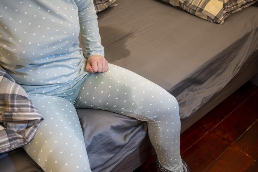 Frau mit Inkontinenzproblem sitzt im Bett - Inkontinenz-Matratzenauflagen helfen