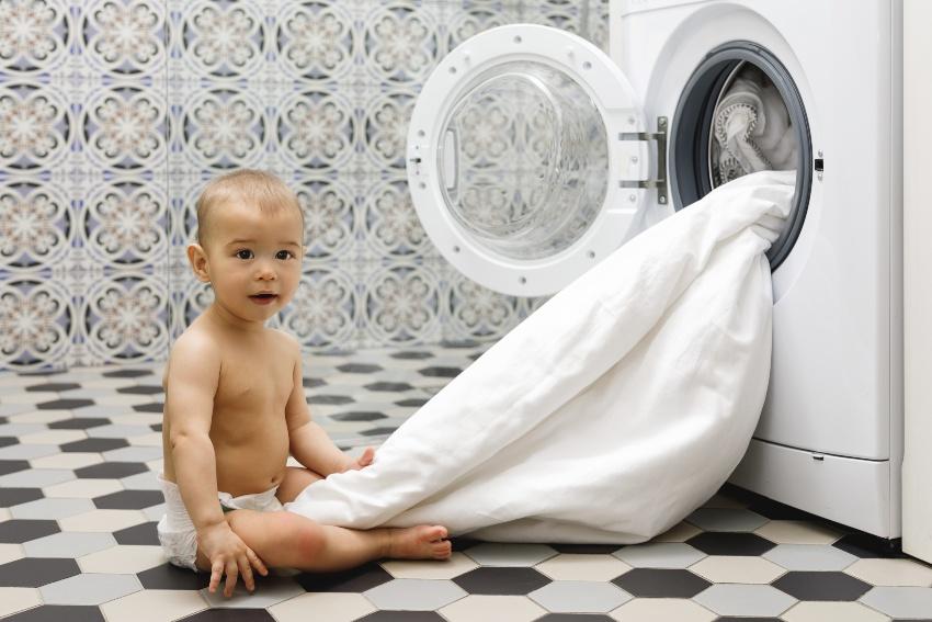 Baby sitzt vor Waschmaschine aus der eine Bettdecke hängt - Bettdecke im Winter