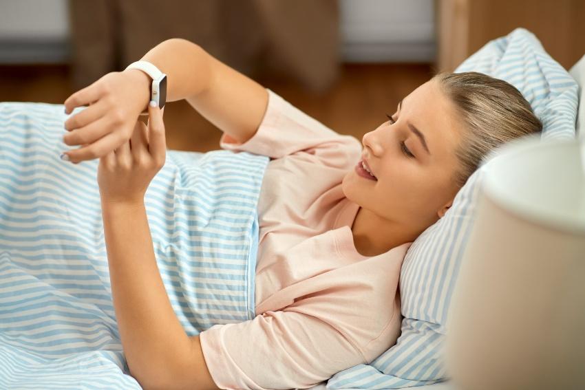 Junge Frau sieht im Bett liegend auf ihre Armbanduhr