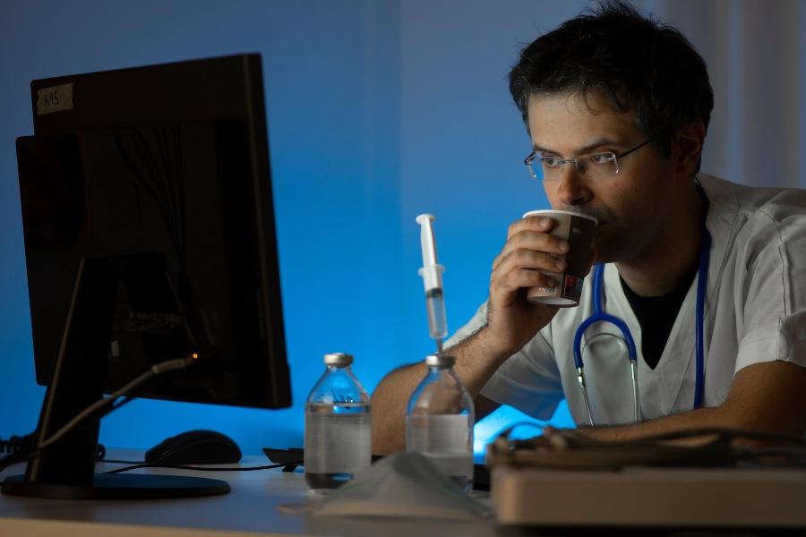 Junger Arzt oder Pfleger sitzt während Nachtschicht am Computer und trinkt Kaffee - Besser schlafen nach der Nachtschicht