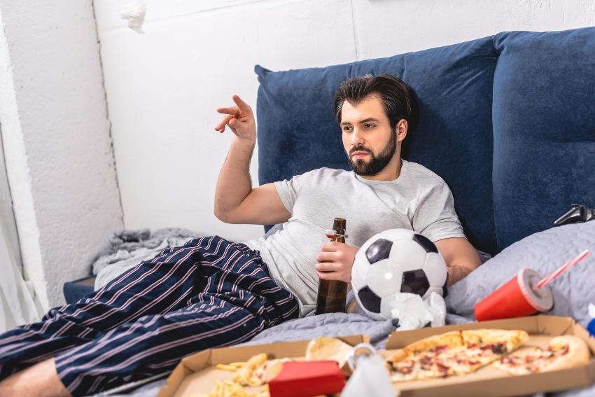 Junger Mann im Bett, Bierflasche in der Hand, Pizza liegt griffbereit